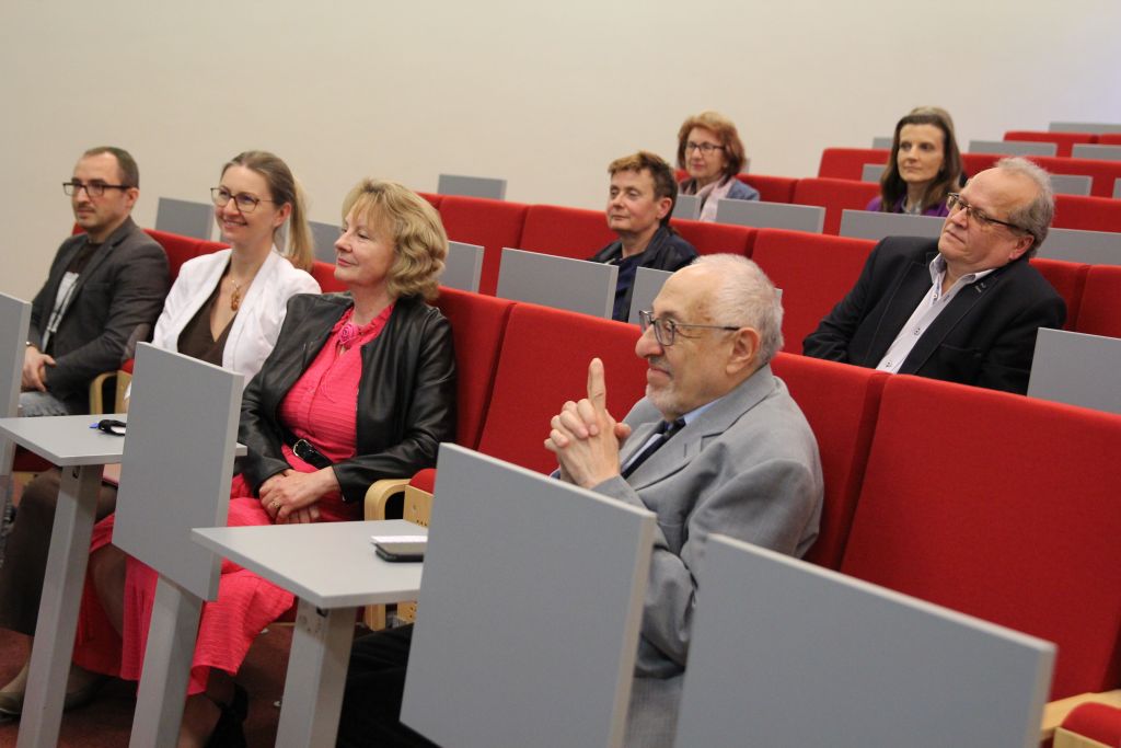 Uczestnicy spotkania siedzą na czerwonych fotelach w auli