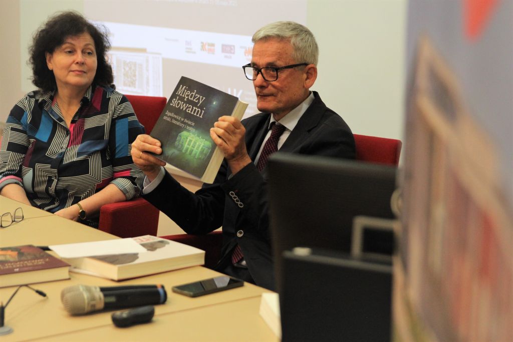 Mężczyzna w okularach i garniturze prezentuje książkę 'Między Słowami'. Obok siedzi kobieta. Przed nimi leżą egzemplarze książek i mikrofony
