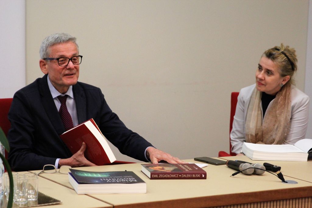 Mężczyzna w okularach i garniturze, trzymający książkę, przemawia do uczestników. Po prawej siedzi kobieta