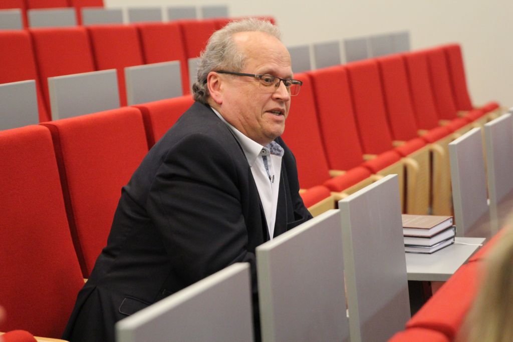 Mężczyzna w okularach i ciemnej marynarce siedzący na czerwonym fotelu w auli, zabrał głos