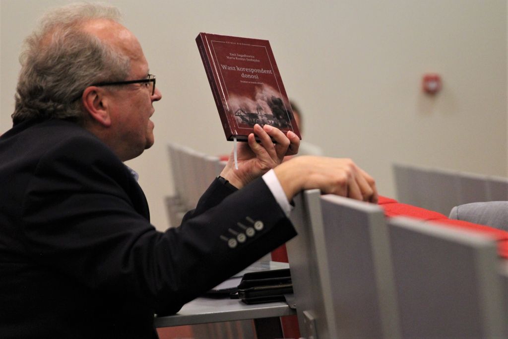 Mężczyzna w okularach i ciemnej marynarce, siedzący w auli, prezentuje książkę z czerwoną okładką zatytułowaną 'Wasz korespondent donosi'