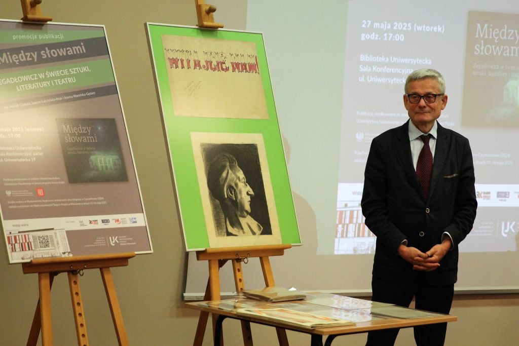 Mężczyzna w okularach i garniturze stoi obok dwóch plakatów promujących spotkanie