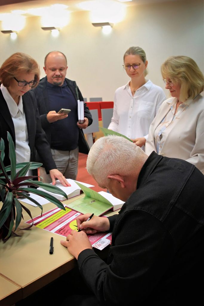 Autor rozdaje autograf przy plakacie a w tle uczestnicy spotkania