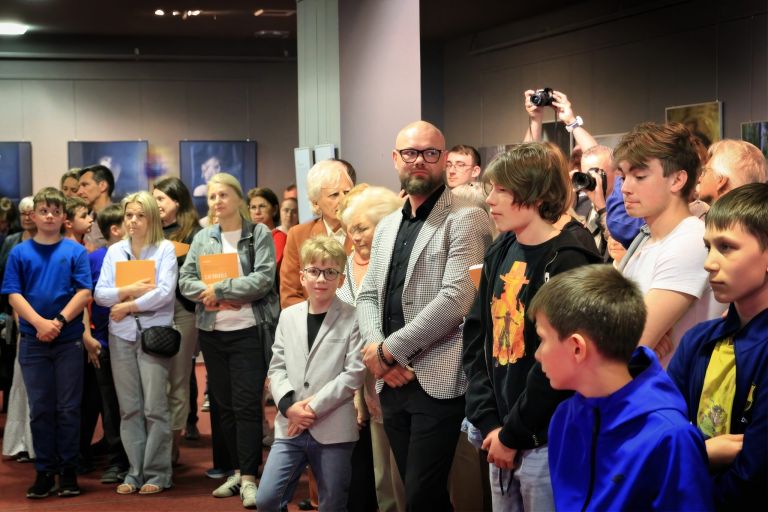 Osoby uczestniczące w wernisażu stoją przed mikrofonem podczas przemówień.